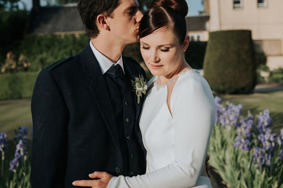 Groom kisses brides head as she looks down in gardens of their wedding venue, brides makeup by Jennifer Louise Studio.